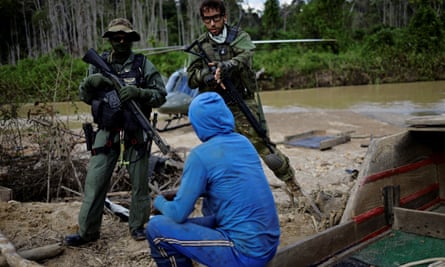 Two men in military-style uniforms, standing holding rifles, talk with a man sitting on a log