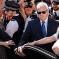 Huw Edwards, in sunglasses and a suit and tie, holds the edge of a car door as he stands, frowning, in a throng of police officers