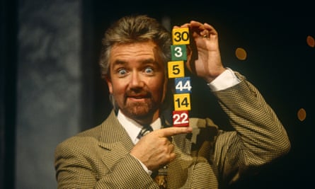 Noel Edmonds in a jacket and tie holding up a stack of lottery numbers on cubes