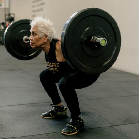 A woman with short, white hair wearing black leggings and vest top, with her knees bent, lifting a large dumbbell in a gym