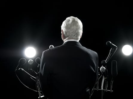 A man with grey hair in a suit stands in front of a microphone with his back to the camera
