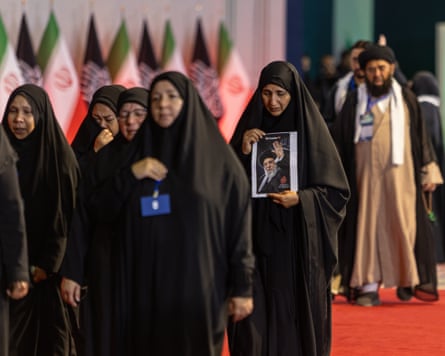 A group of women, some weeping, wearing black chadors walk past the coffins