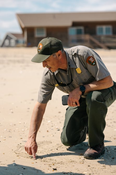 A man in a National Park Service uniform draws a line in the sand with a home behind him on the beach