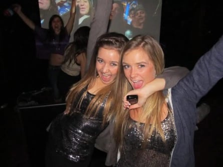 Lauren holding her phone in her left hand, with her arm draped around Drew’s neck, both wearing sequin dresses and laughing while looking at the camera