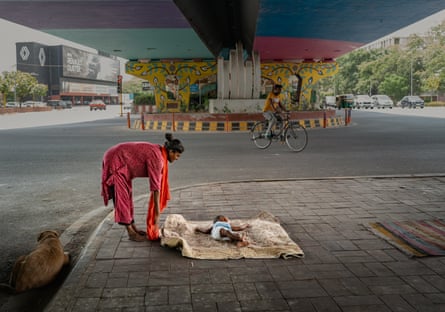 Shahida bends over baby Jannat, who is lying on a blanket on the pavement near a busy road as buses and cars pass