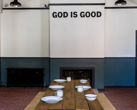 Tin plates and cups laid out on a long wooden table under the words ‘God is good’ painted on the wall