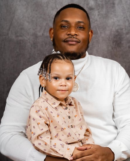 A man in a white turtleneck hugs his daughter in a posed portrait against a gray backdrop.