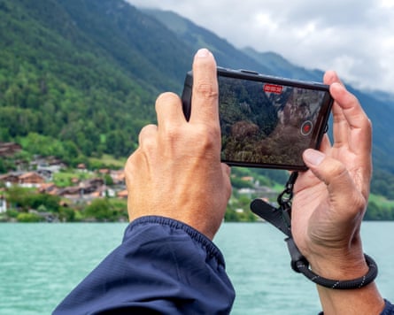 Tourist’s hands holding smartphone, taking a photo or a video of a village on a green mountain near a lake on a sunny day