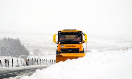 A yellow gritting lorry with snow plough pushes snow towards the side of a road in a white, snowy landscape