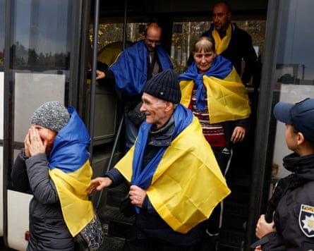 Evacuated residents wrapped in Ukrainian flags exit a bus as a police officer looks on