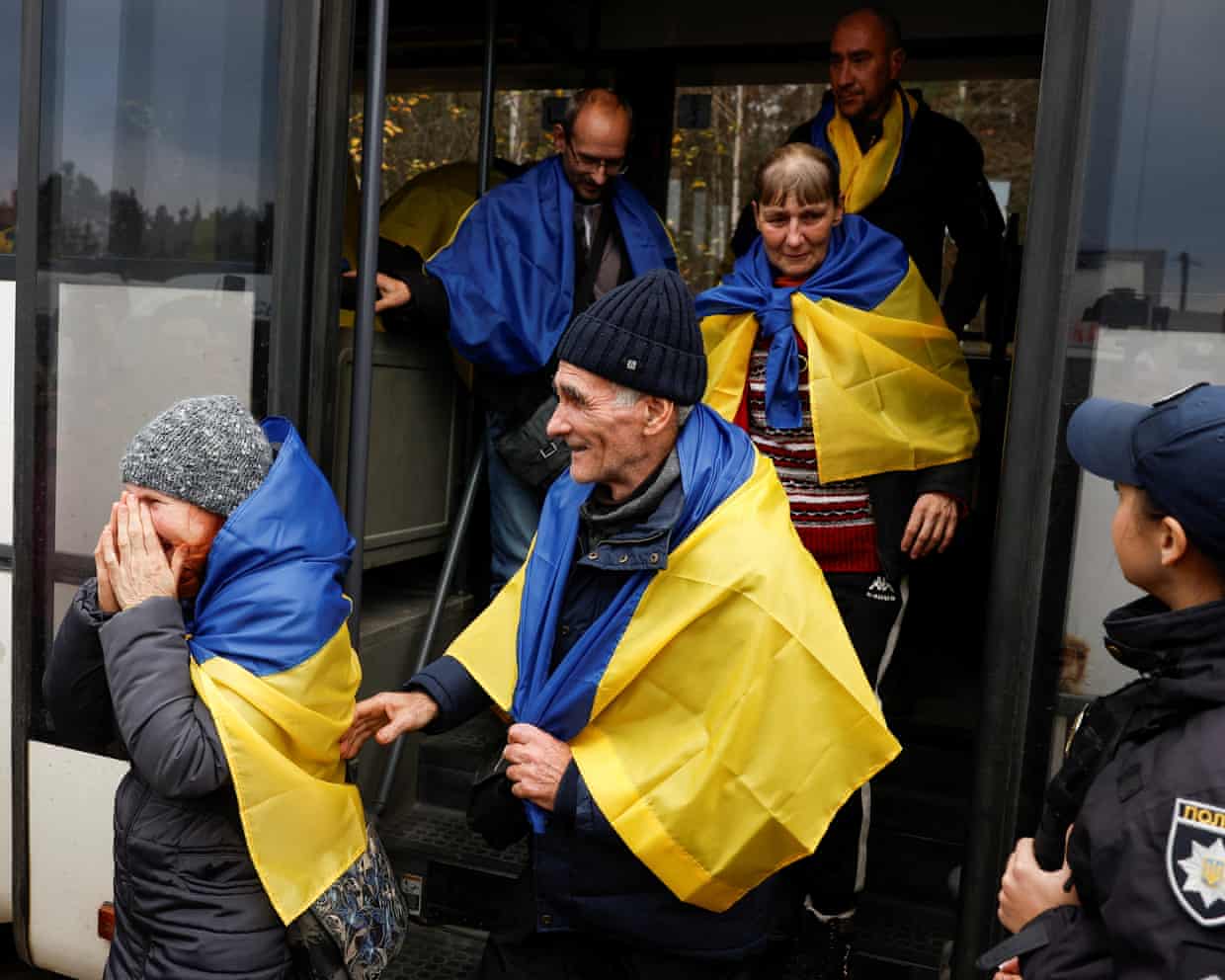 Evacuated residents wrapped in Ukrainian flags exit a bus as a police officer looks on