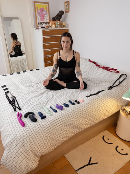 Portrait of Carmen sat cross legged on a bed with her sex toys arranged in a square around her