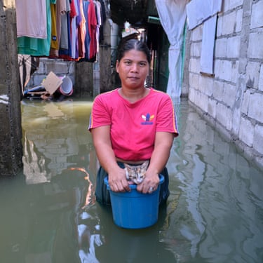 A woman stands thigh deep in floodwater holding a bucket with a cat in it, looking out over the edge