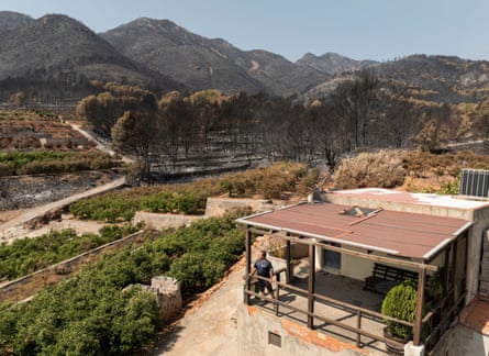 A man at the entrance of his low-rise house in a dry, mountainous landscape, with greenery around it but blackened and burnt trees in the background.