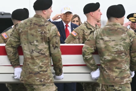 Donald Trump saluting while wearing a Trump baseball cap as coffins of US military personnel are carried past.