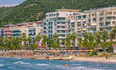 A beach in Vlora, Albania, with people in the sea, on the beach and with buildings and mountains in the background.