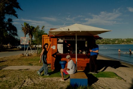 A man sits under a parasol outside a wooden hut next to a sandy beach