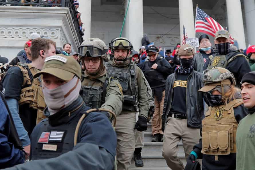 A group of men and women in military fatigues march down the steps of the Capitol in formation amid a mob of people waving American flags.