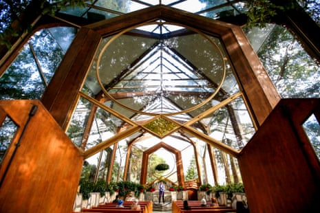 people attend a church service in a wood structure that is mostly glass and looks up at green canopies of trees.