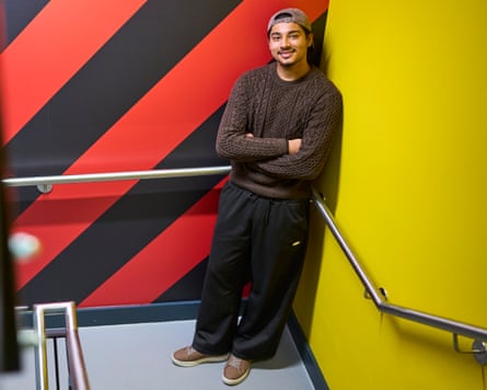 Hamid stands in a corner on a stairway, smiling. The walls behind him are painted bright yellow on one side and with red and black diagonal stripes on the other. He wears baggy black trousers and a brown cable-knit jumper, plus a backwards-facing baseball cap.