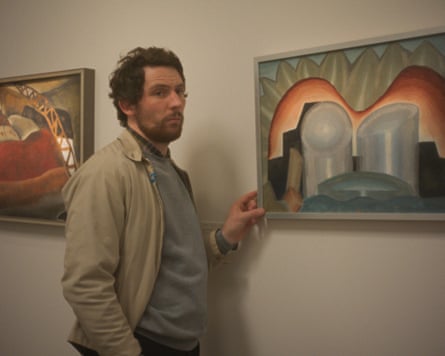 Josh O’Connor stands in a gallery with his hand on the frame of a painting.