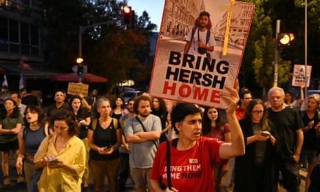 A woman in a red T-shirt with the words Bring Them Home Now across it and holds a placard reading Bring Hersh Home under a picture of the hostage; a crowd of people is gathered behind her, some others with placards and signs