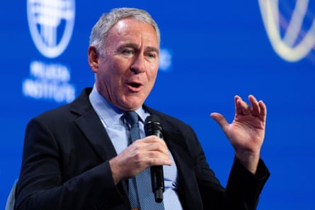 A man in a black suit, light-blue shirt and blue tie, with grey hair, speaking into a microphone