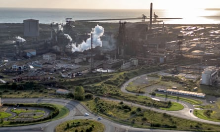 An overhead drone view of the Tata steelworks in Port Talbot showing the site from a distance across roads and green areas and with the sea in the background; the sun is shining across the water giving a gentle glow