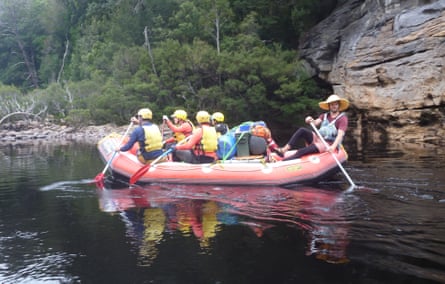 A group of rafters wearing yellow helmet and lifejackets, and holding red paddled oars, sitting in a raft on the Franklin river in Tasmania.