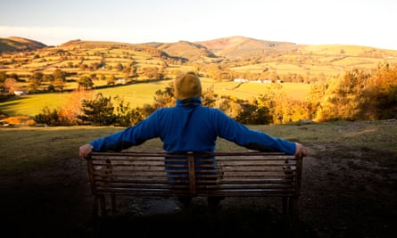 Back view of a hill walker sitting on a bench, his arms spread across the back of it, looking towards the Clwydian Range bathed in early morning light