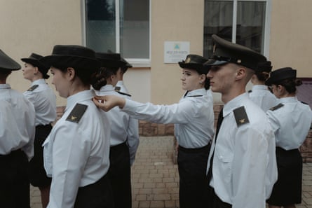 One cadet straightens her classmate’s collar. Male and female cadets line up in front of a pale cream building. They all wear white shirts with epaulettes and black peaked caps.