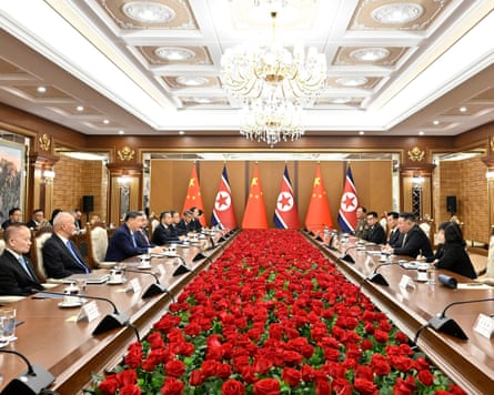 Xi Jinping and Kim Jong-un face each other across a long table with red flowers surrounded by officials