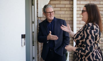 Keir Starmer stands outside a white front door with Labour's candidate for Stafford, Leigh Ingham. He wears glasses, a dark shirt and dark suit, while she is seen standing side-on with a hand raised as she talks; she is in a cream and black patterned dress and they are both seen standing in front of a pale brick wall.