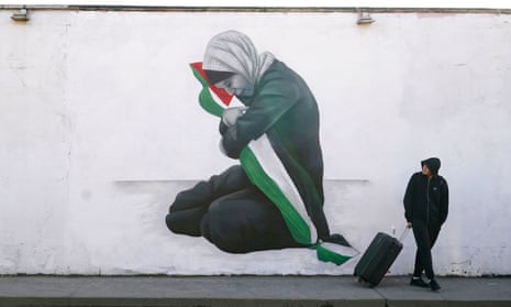 A man with a suitcase and hoodie looks at a mural by the artist Emmalene Blake of a girl wearing a headscarf and hugging a Palestinian flag with her head bowed