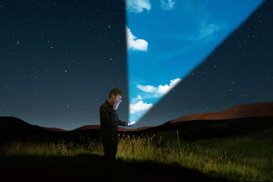 A man standing in a nighttime landscape looking at his phone, which is illuminating a sunny, bright beam into the sky.