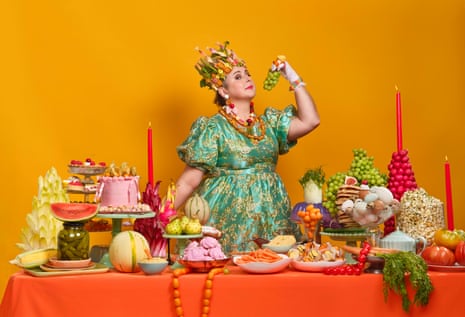 Siobhan McSweeney in a floral headdress and green gown holds grapes beside a table of food