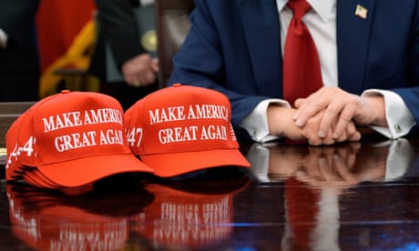 Two red baseball caps with the words 'Make America great again' on them sit on a table. A man's folded hands can be seen behind them.