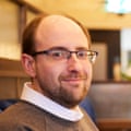 A young, balding man with glasses, wearing a grey top over a white shirt