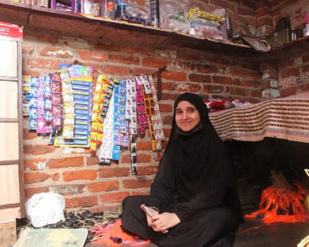 A young woman in a black veil sits in a brick room, with goods in colourful packaging hanging next to her