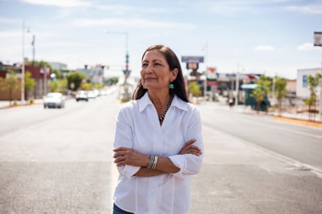 Deb Haaland stands on a street with crossed arms, wearing turquoise jewellery, looking into the distance
