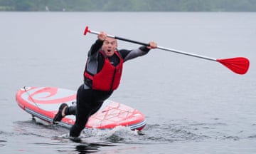 Ed Davey wearing lifejacket and holding a long paddle looks at camera as he 'falls' off a paddleboard on water