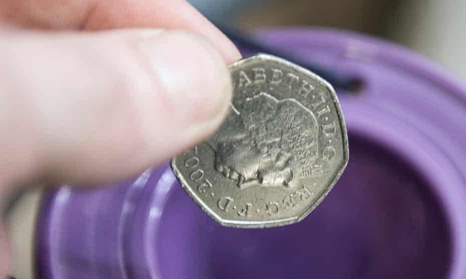 A very close-up photo of 50p coin being dropped into a purple charity box by thumb and forefinger