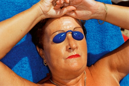 Close -up of the face of a woman lying in the sun, head on a blue towel, hands resting at the top of her forehead, wearing blue goggles and red lipstick