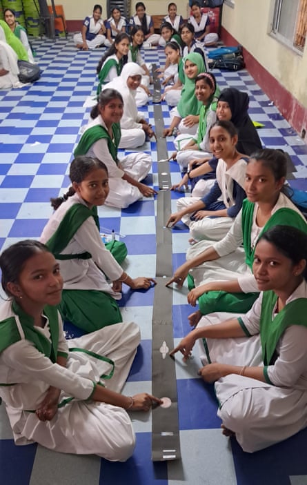 Female schoolchildren sit cross legged in two lines, either side of a long strip of black paper with white images of planets on it