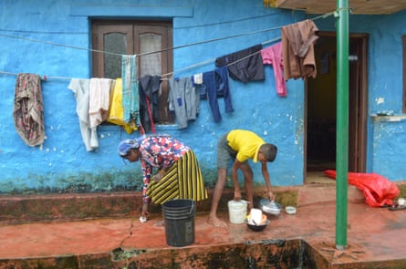 A woman with her son, who is washing dishes, outside a small blue-painted house with a string of laundry on a line 