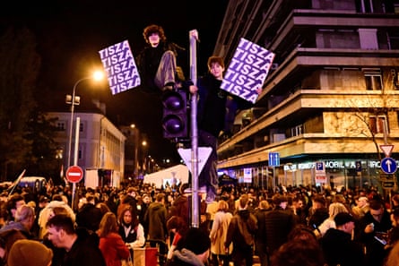 Two boys climb up a lamp post to hold up signs that say Tisza on a crowded street in Budapest
