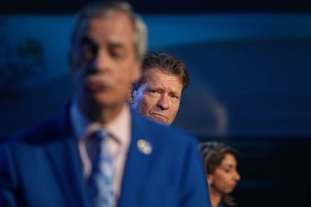 Richard Tice looking at Nigel Farage during the press conference with Suella Braverman in the background.