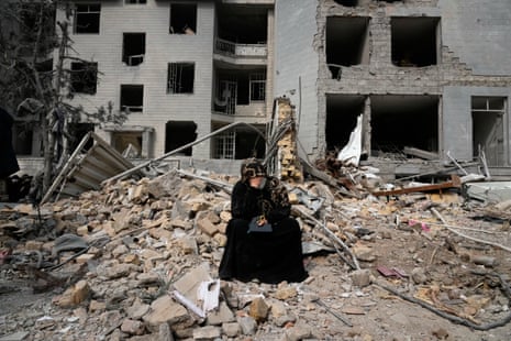 A woman in a black dress and a black hijab cries into her hand as she sits on rubble and debris in front of the grey shell of a building.