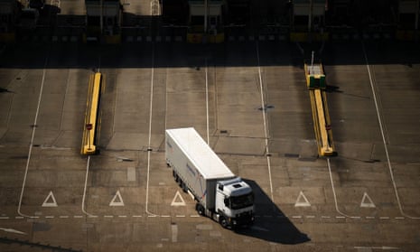 A white lorry passes through a checkpoint at the Port of Dover: it is seen from above on an expanse of grey tarmac with marked-out lanes, having emerged from a strip of dark shadow