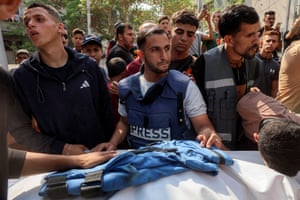 A group of men gather around a white body bag with a blue bulletproof press vest laid on top